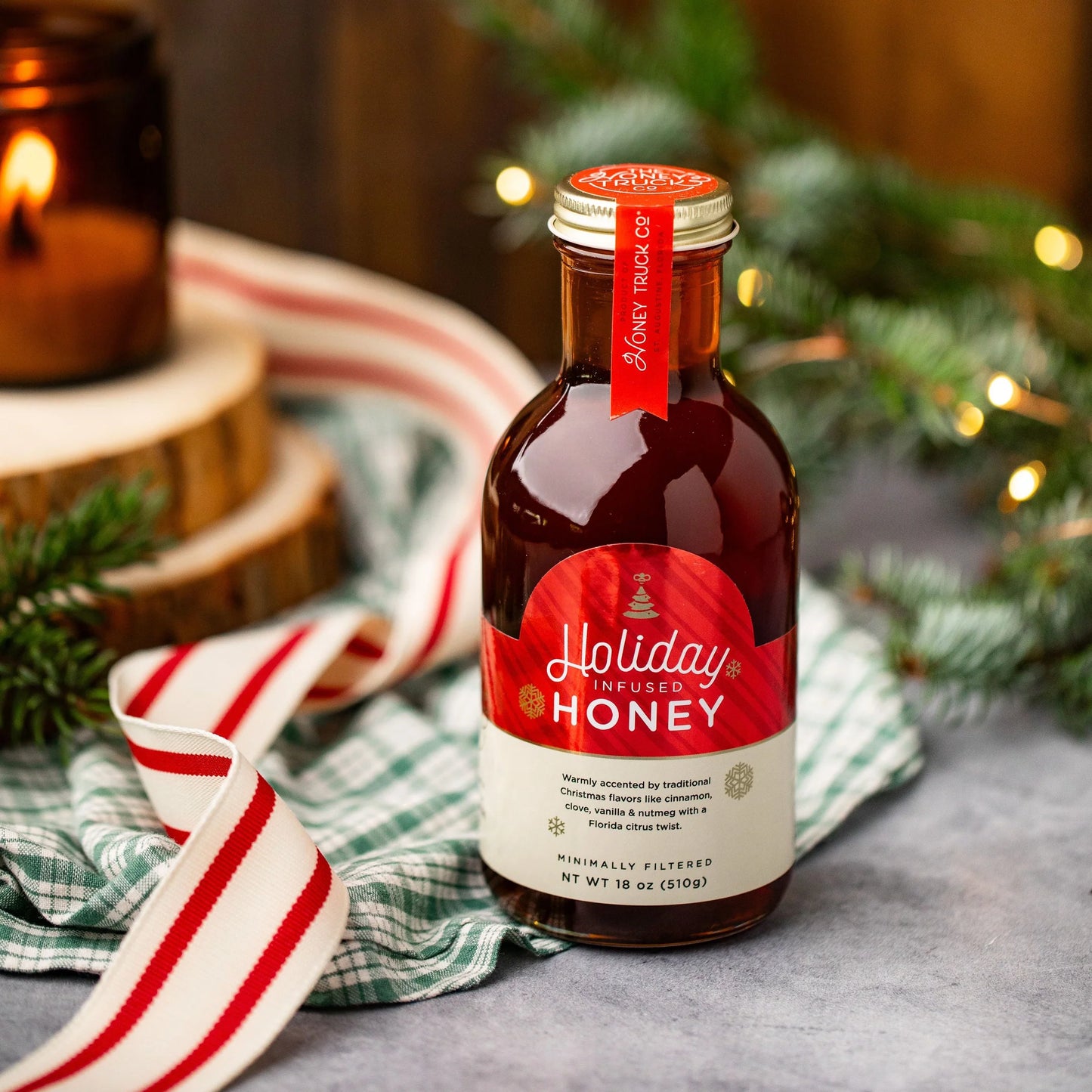 A glass bottle of infused honey with ribbon, wood, fir boughs, candles and holiday lights in the background