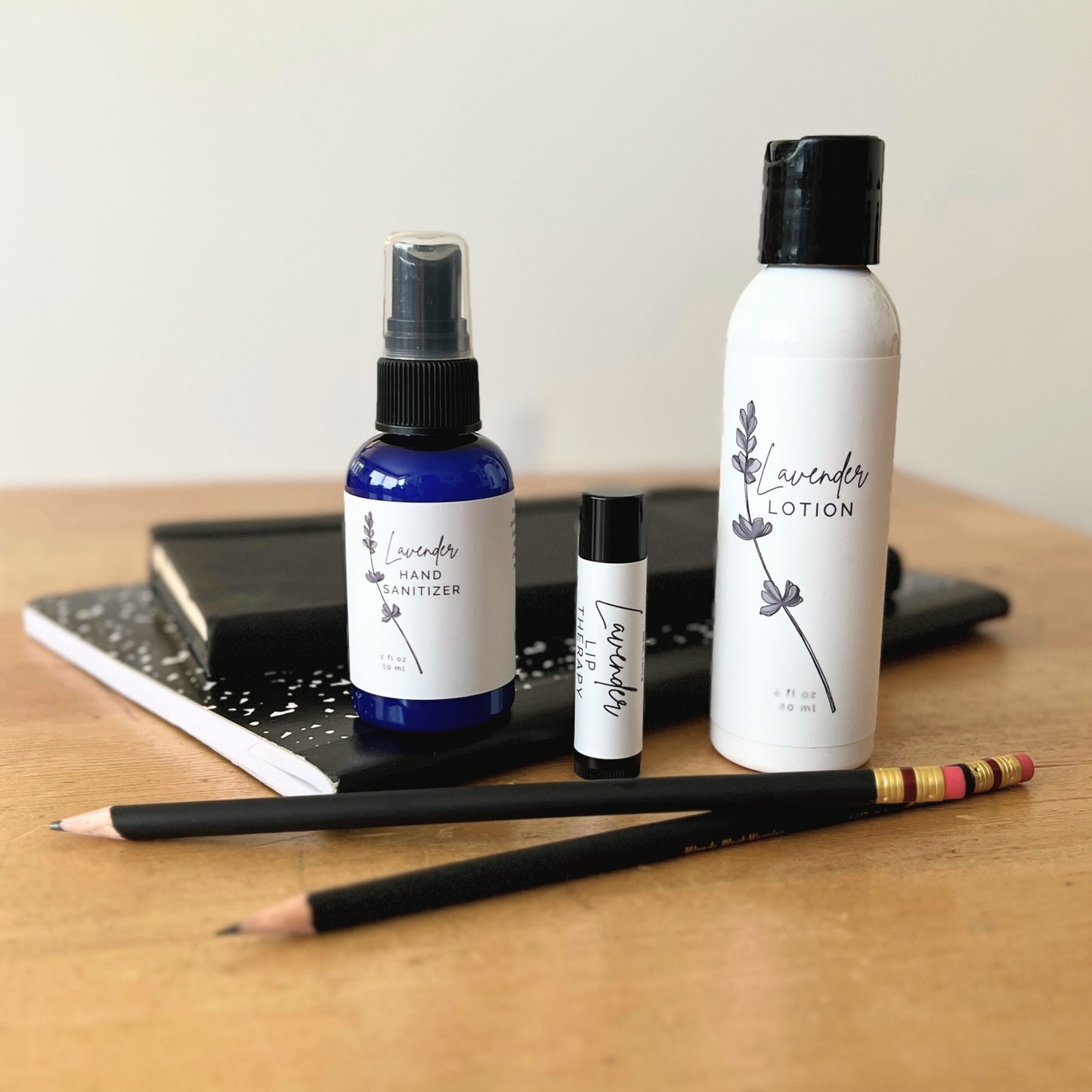 Three bottles of lavender products including lotion, hand sanitizer, and a small bottle on a wooden surface with a notebook and pencils.