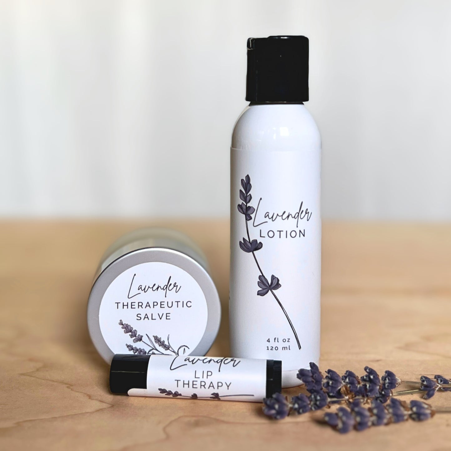 Bottle of lavender lotion, black tube of lavender lip therapy and jar of lavender salve with lavender flowers on a wood and white background