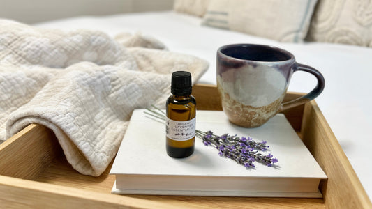 A bottle of Organic Lavender Essential Oil displayed on a wooden tray with a white bound book, a purple tea cup, purple lavender flowers and a white blanket on a white bedspread.