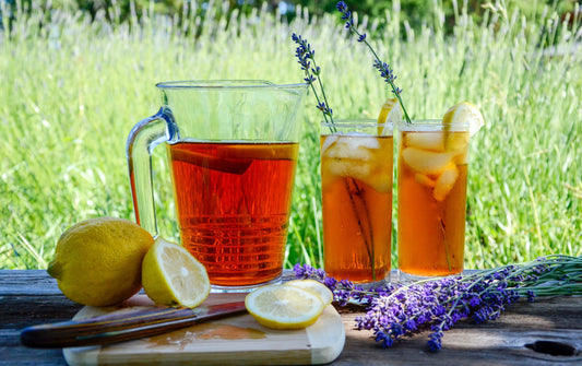 Two glasses of iced tea with lemon slices and a pitcher on a wooden surface with lavender in the background.