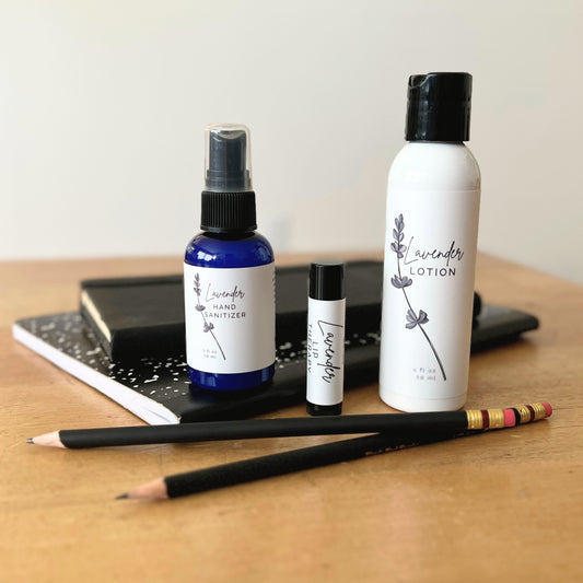 Three bottles of lavender products including lotion, hand sanitizer, and a small bottle on a wooden surface with a notebook and pencils.