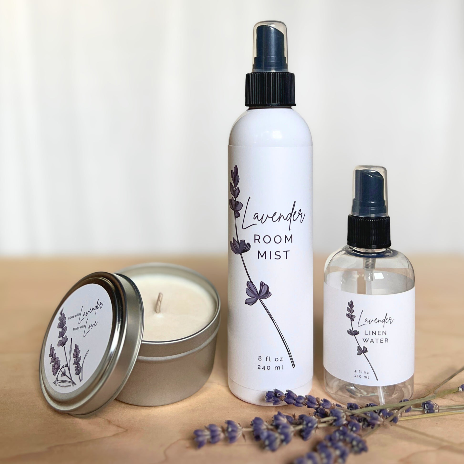 Lavender candle, bottle of lavender linen water and bottle of room mist with lavender flowers on a wood and white background