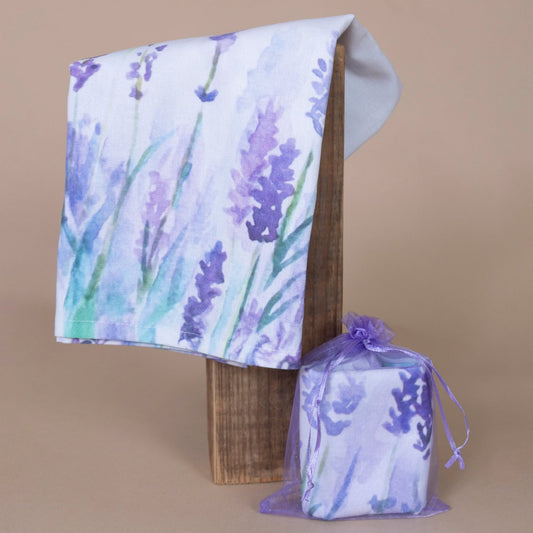 Lavender Tea Towel hanging on display and elegantly packaged in a small purple bag against a beige background.